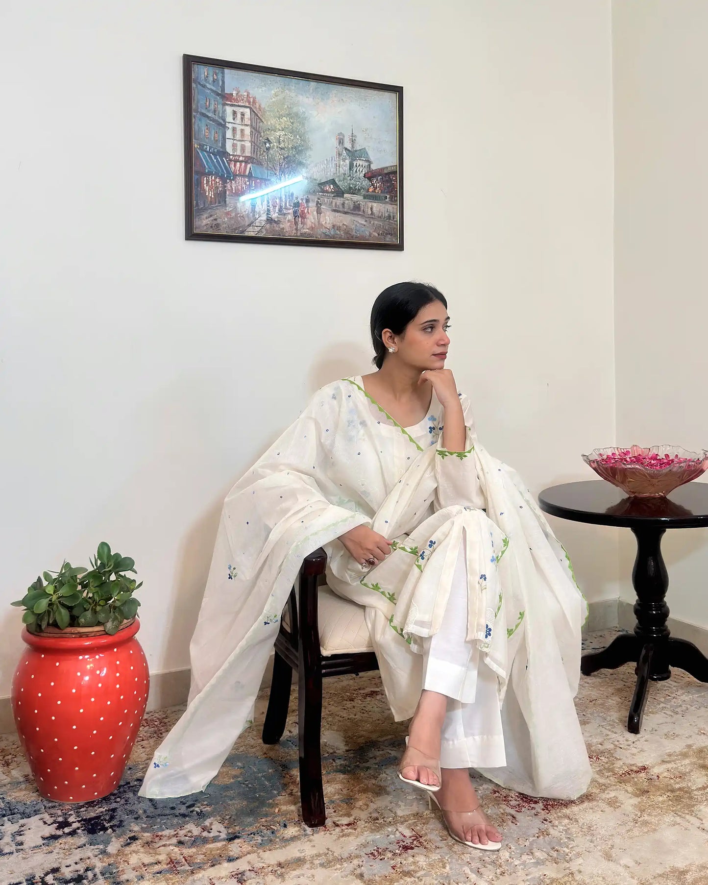 Model seated in a chair with chin resting on hand, looking to the side, wearing the off-white chanderi kurta and dupatta with green appliqué trim visible along the front panel.
