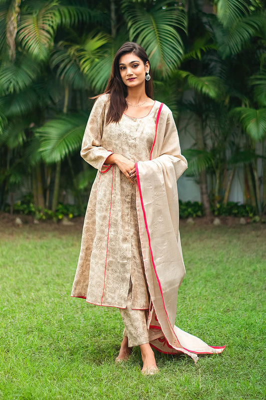 An indian woman wearing a printed staple tussar gold A line kurta and trousers with gold chanderi dupatta. The dupatta has red calligraphy motifs appliqued at the borders of  the dupatta. The woman looks straight in the camera