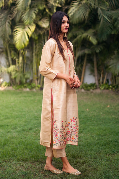 Side view of an Indian lady dressed in a beige silk kurta with resham floral border and beige trousers.