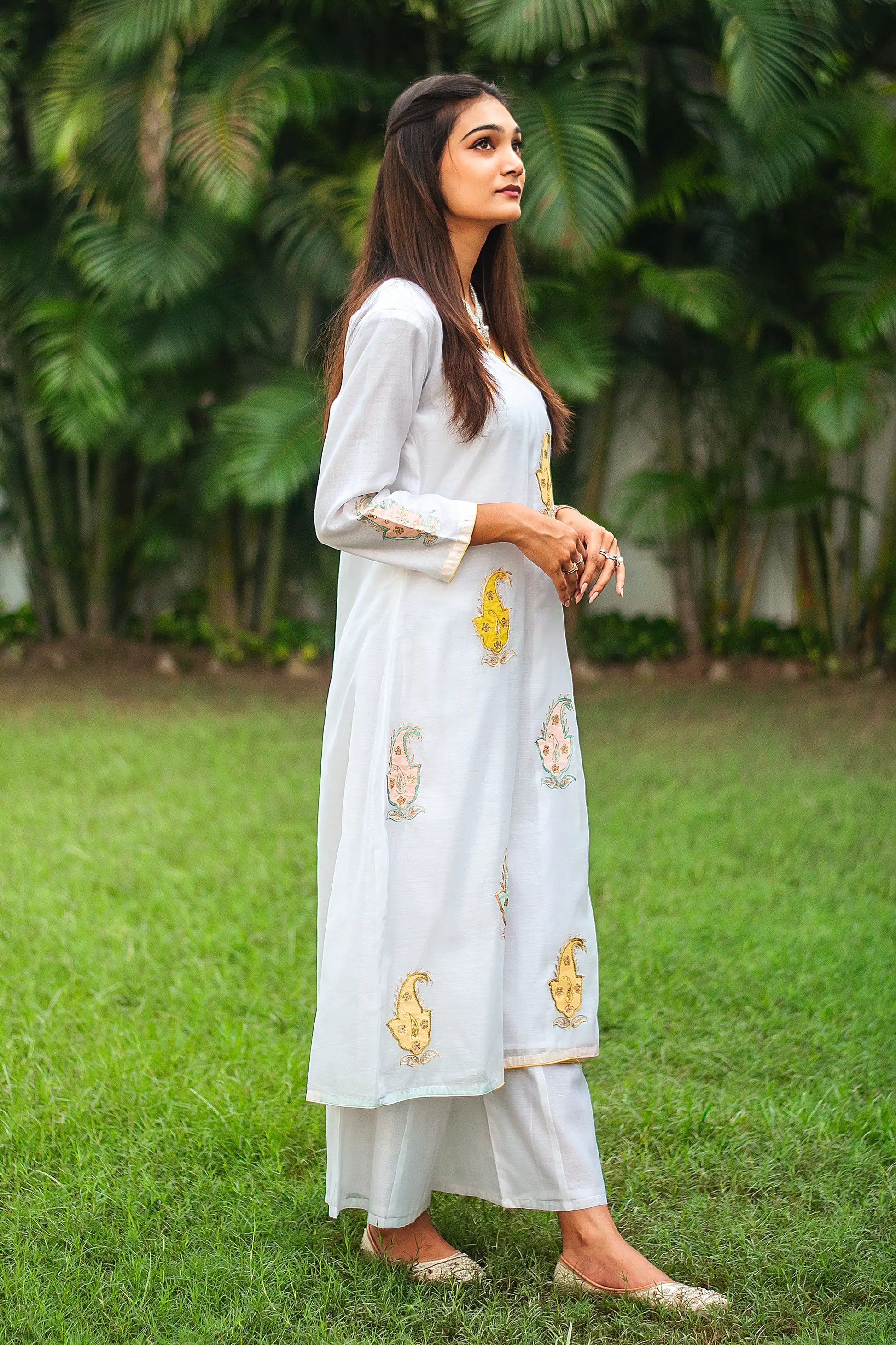 Side pose of an Indian woman wearing a white chanderi angarkha kurta with kairi patchwork, complemented by white palazzo.