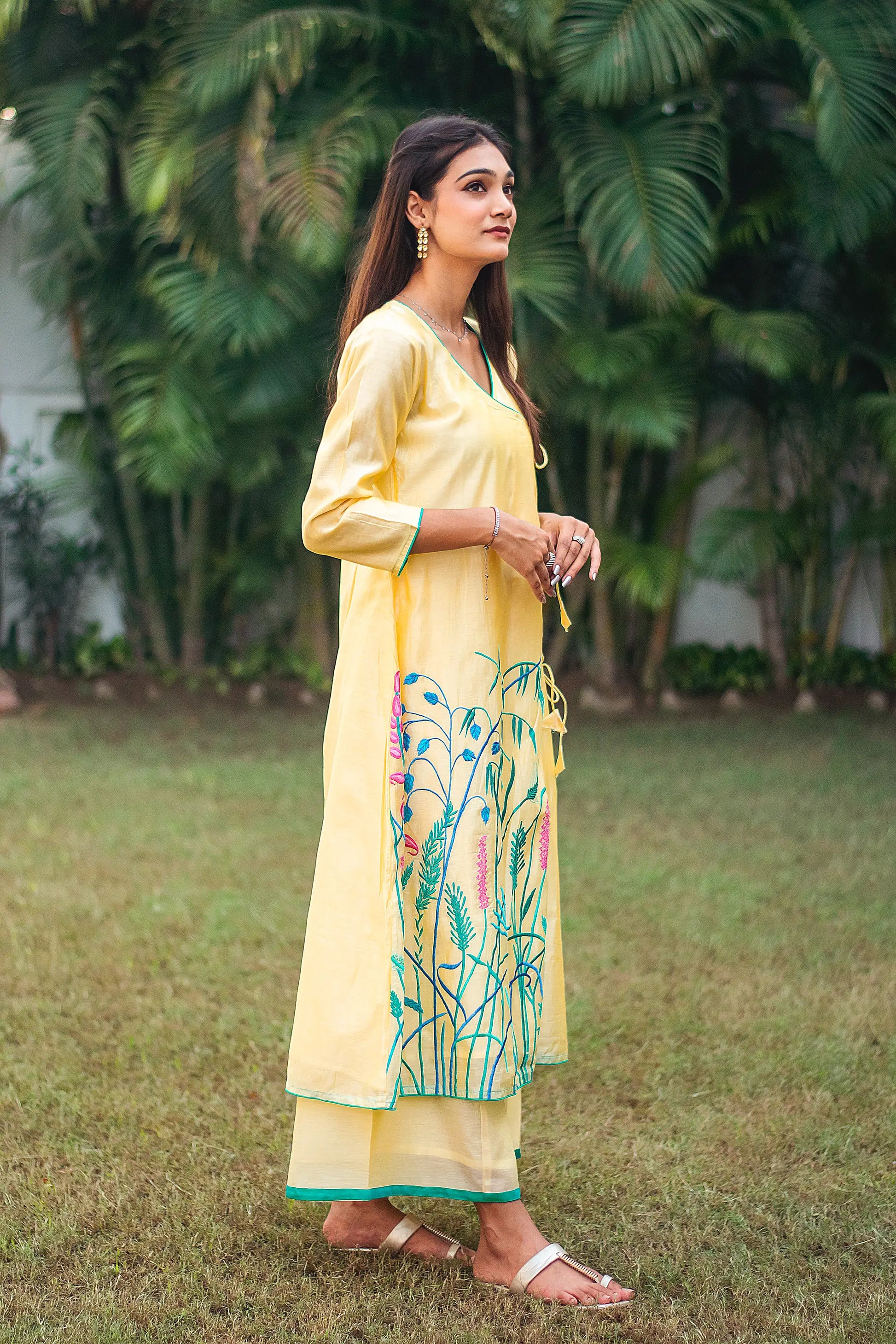 Side pose of an Indian model wearing the yellow angrakha kurta with pink and teal embroidery, paired with yellow palazzo.