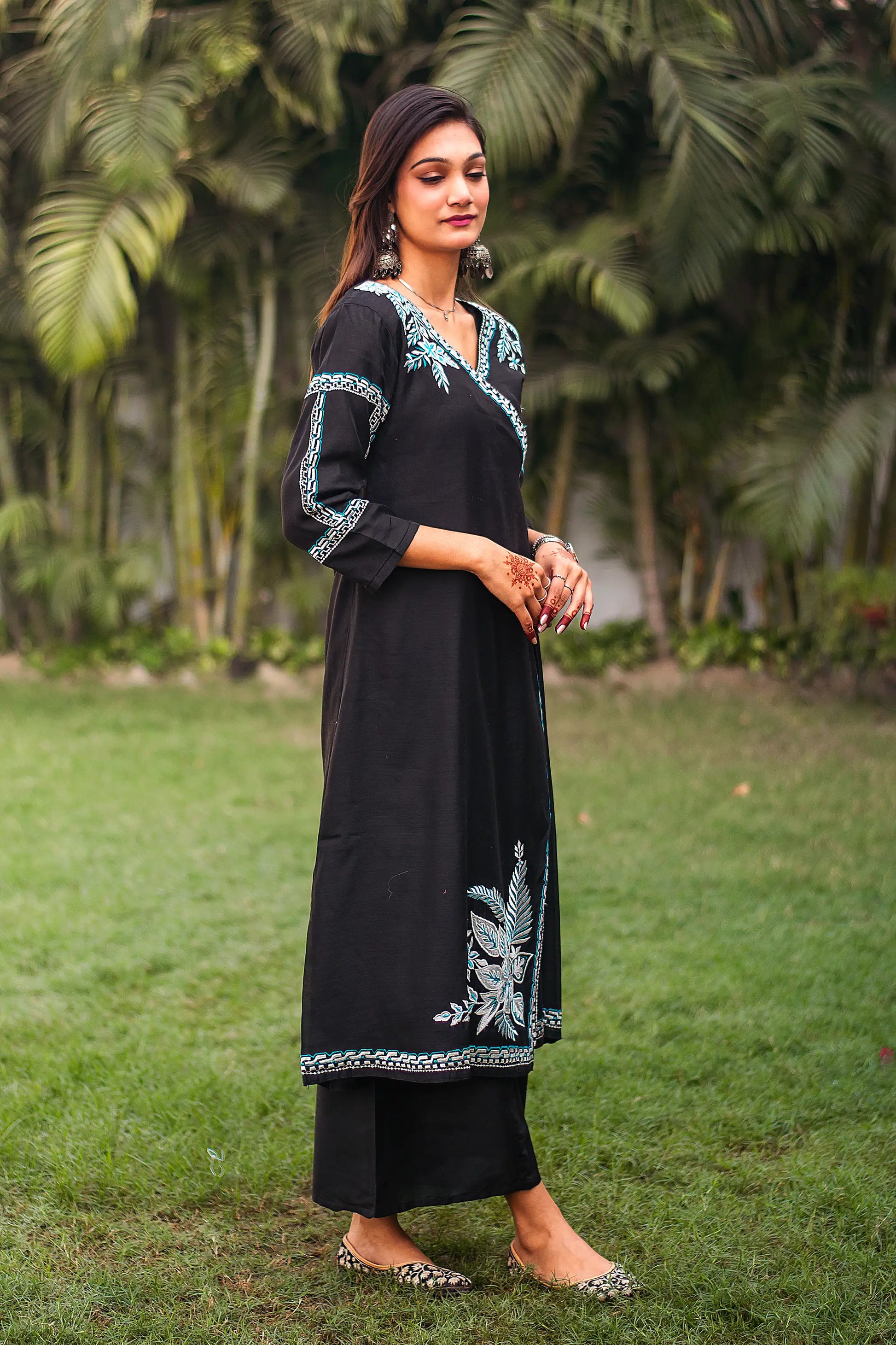 An Indian woman gives a side pose in a black chanderi angrakha kurta with white and blue embroidery, paired with black palazzo.