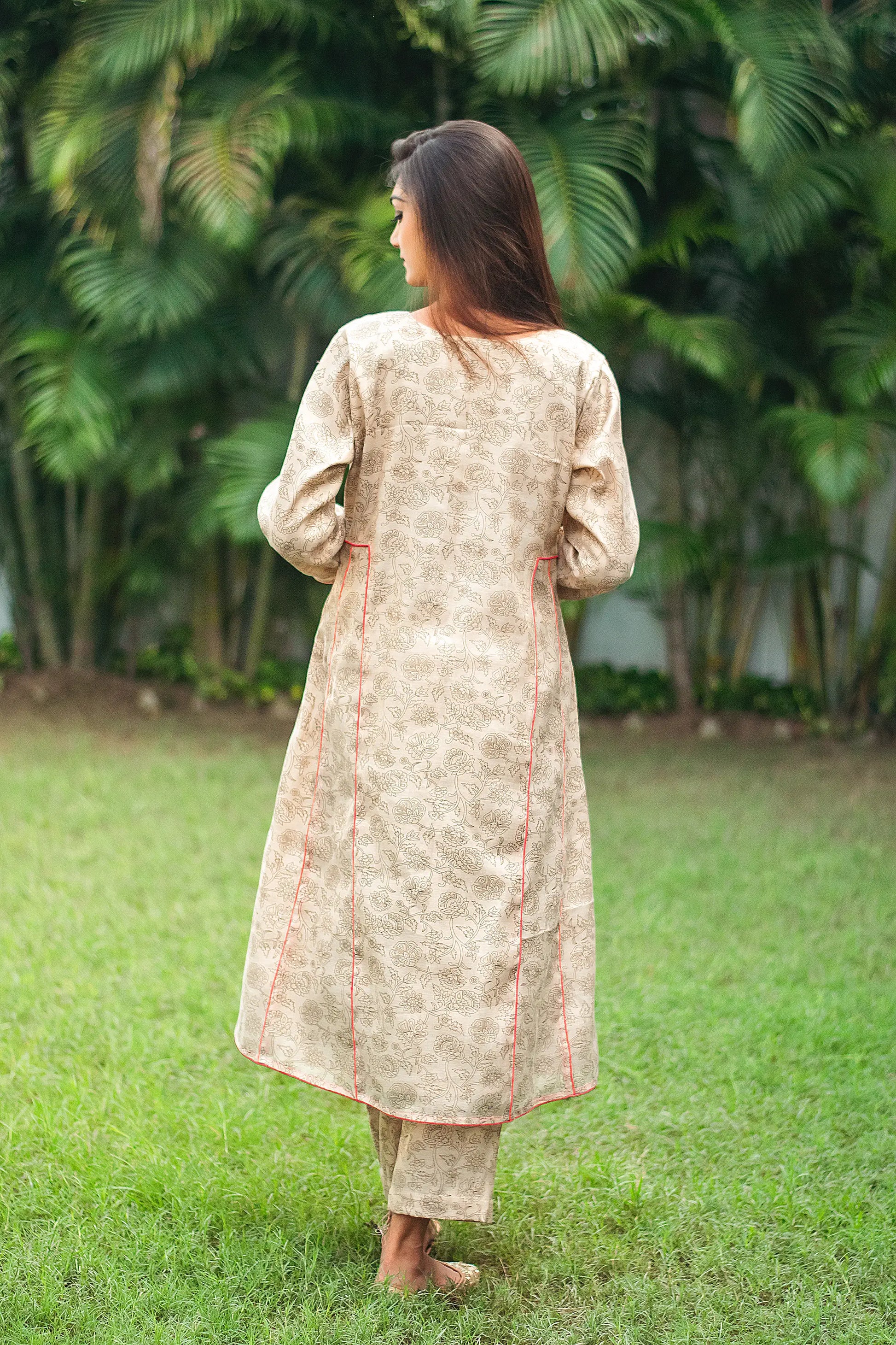 An indian woman wearing a printed staple tussar gold A line kurta and trousers. An indian woman wearing a printed staple tussar gold A line kurta and trousers with gold chanderi dupatta. The A line kurta has double kali in red colour.The woman is standing with her back towards the camera and showcases the A line kurta from the back.