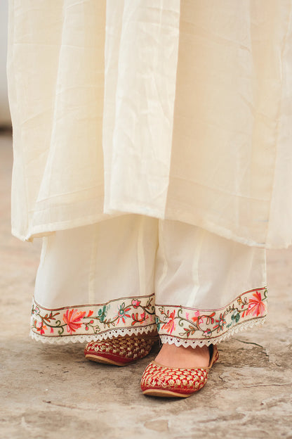 Close-up of the off-white palazzo pants and embroidered juttis of a young Indian woman, showcasing the intricate Kashmiri embroidery on the hems.