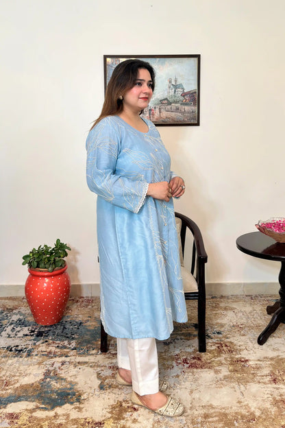 Side view of the blue chanderi A-line kurta with white floral resham embroidery, worn with off-white trousers, dupatta off.
