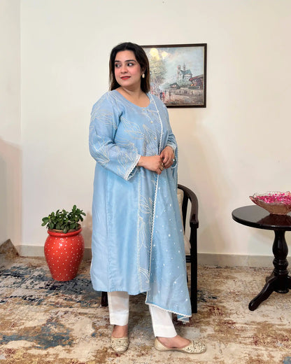 Front view of the blue chanderi A-line kurta with white floral resham embroidery, dupatta off, worn with off-white trousers.
