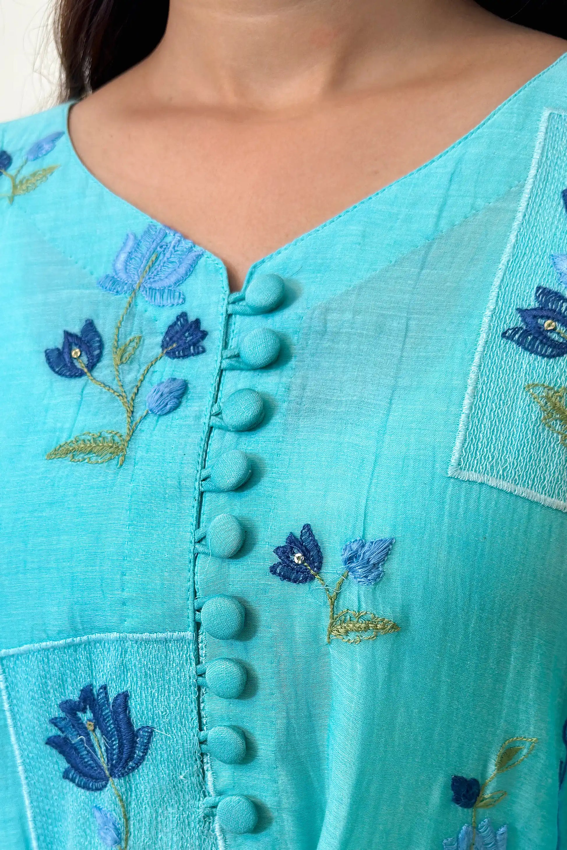 Close-up of the button placket on an aqua-blue chanderi kurta, showing blue floral resham embroidery around the neckline area.