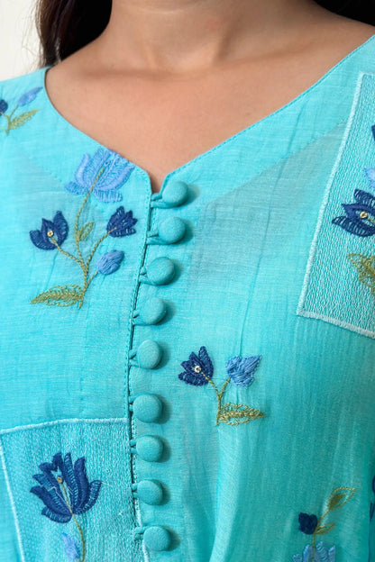 Close-up of the button placket on an aqua-blue chanderi kurta, showing blue floral resham embroidery around the neckline area.
