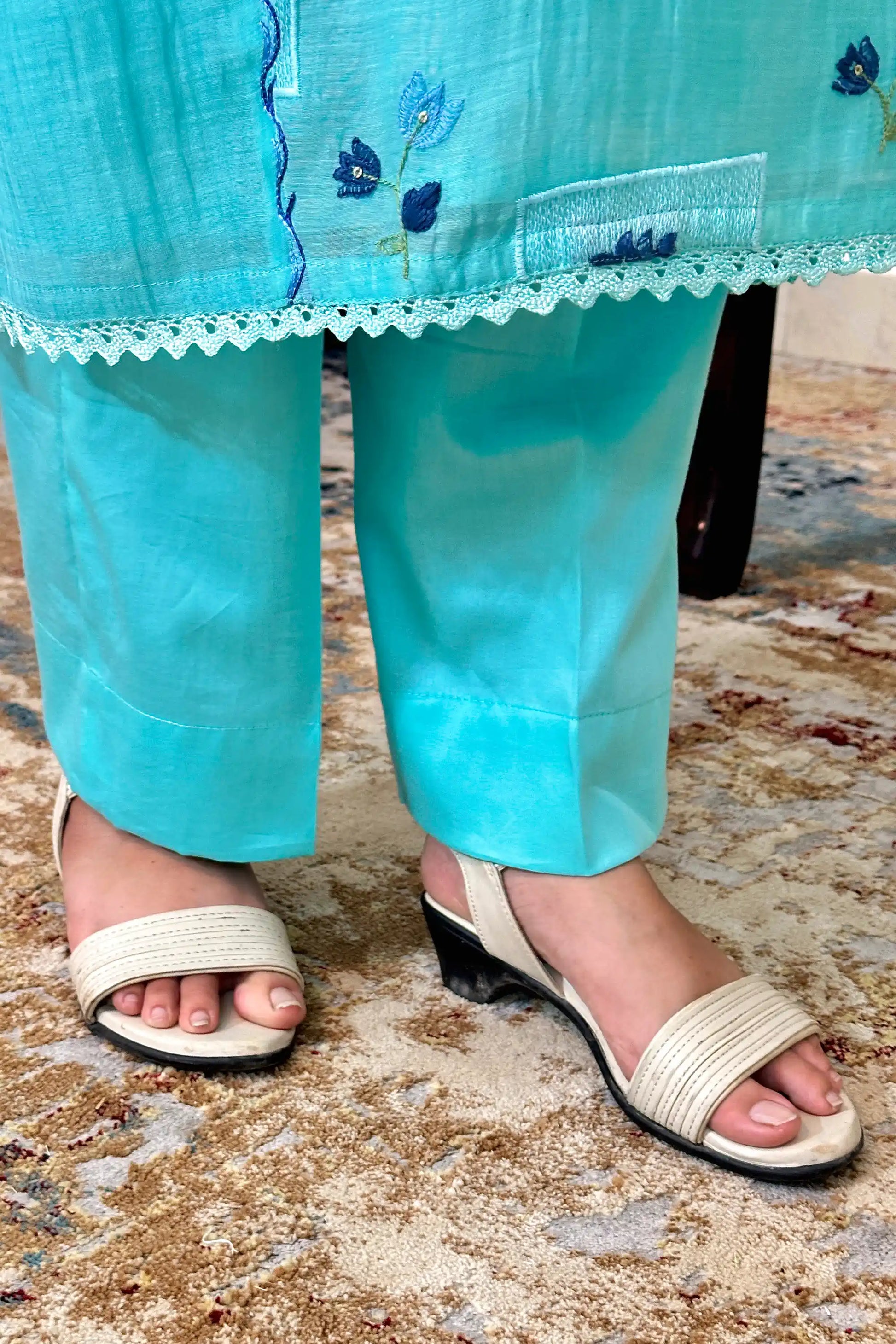 Close-up of the hem and trousers of an aqua-blue chanderi kurta, showing blue floral resham embroidery and a lace-trimmed edge over aqua-blue trousers.