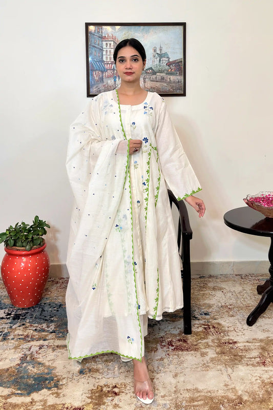 Front view of an off-white chanderi A-line kurta with scattered blue floral embroidery, green appliqué vine trim down the front panels, and a white circular embroidered motif with a blue flower at its center, paired with off-white trousers and a chanderi dupatta with scattered dot embroidery and a green scalloped border.
