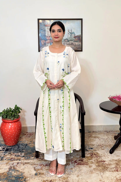 Front view of the off-white chanderi A-line kurta and off-white trousers, showing scattered blue floral embroidery, green appliqué vine trim down the front panels, and a white circular embroidered motif with a blue flower at its center.
