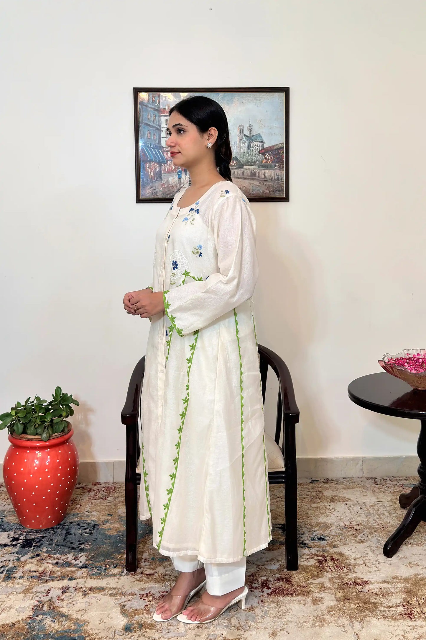 Left side view of the model wearing the off-white chanderi A-line kurta, showing the green appliqué vine trim along the front seam and blue floral embroidery on the shoulder and bodice.