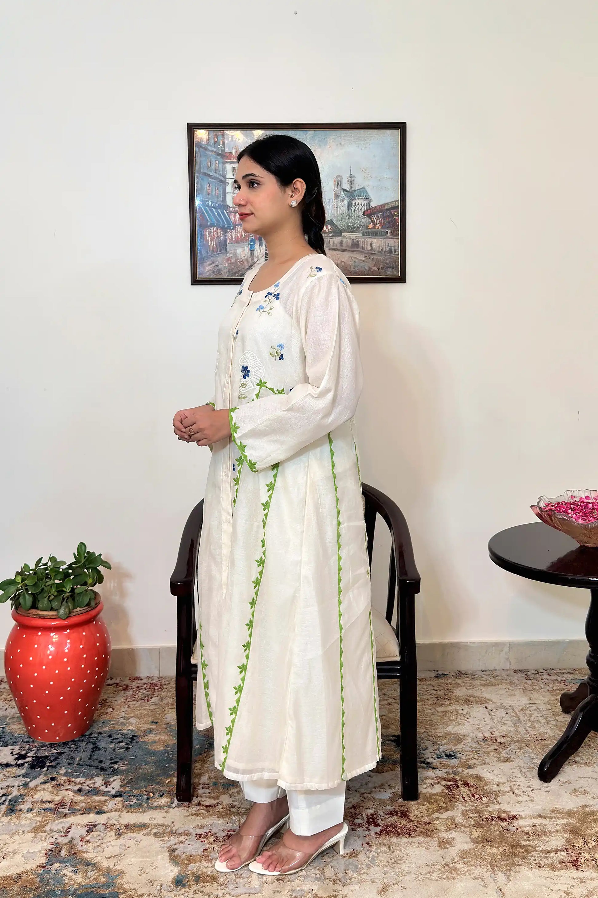 Left side view of the model wearing the off-white chanderi A-line kurta, showing the green appliqué vine trim along the front seam and blue floral embroidery on the shoulder and bodice.