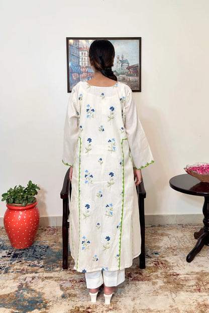 Back view of the model wearing the off-white chanderi A-line kurta, showing scattered blue floral embroidery, green appliqué vine trim down the centre back panel, and a white circular embroidered motif with a blue flower at its center.