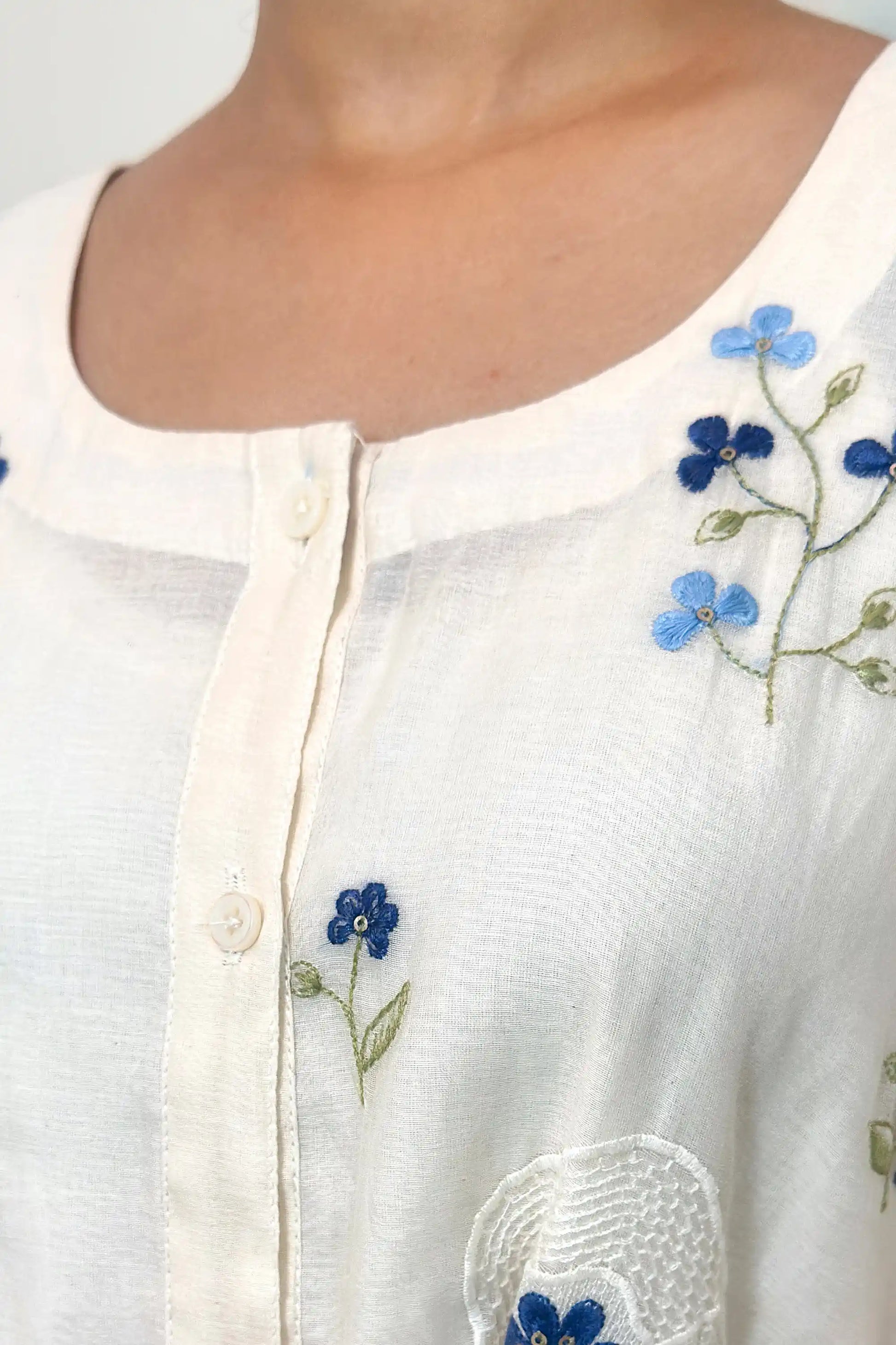 Close-up of the neckline and button placket, showing blue floral embroidery clusters and the edge of a white circular embroidered motif with a blue flower.