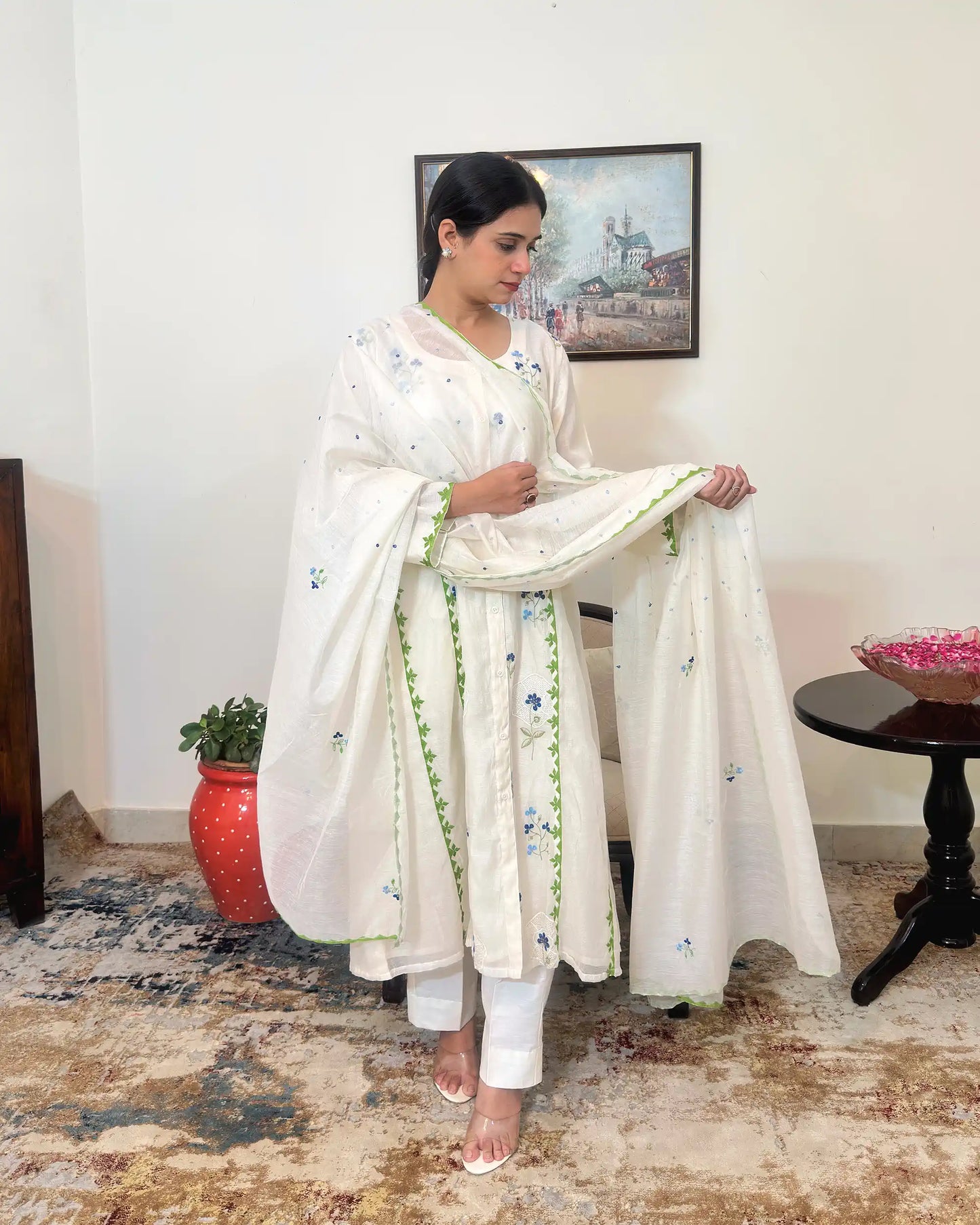 Model standing and holding the off-white chanderi dupatta open with both hands, looking down, showing the scattered dot embroidery and green scalloped border along its length.