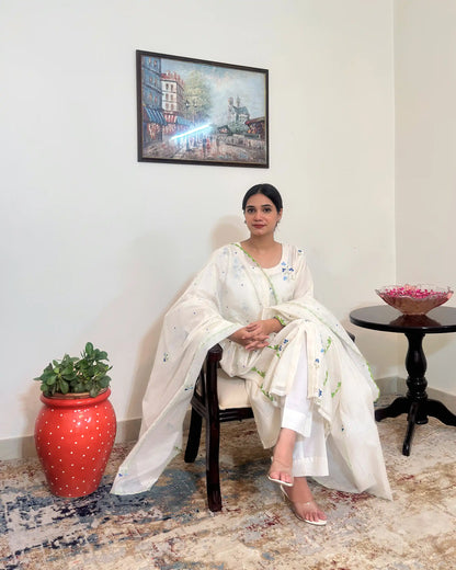 Model seated in a chair, hands folded, wearing the off-white chanderi kurta and dupatta, with the dupatta's scattered dot embroidery and the kurta's green appliqué trim visible.