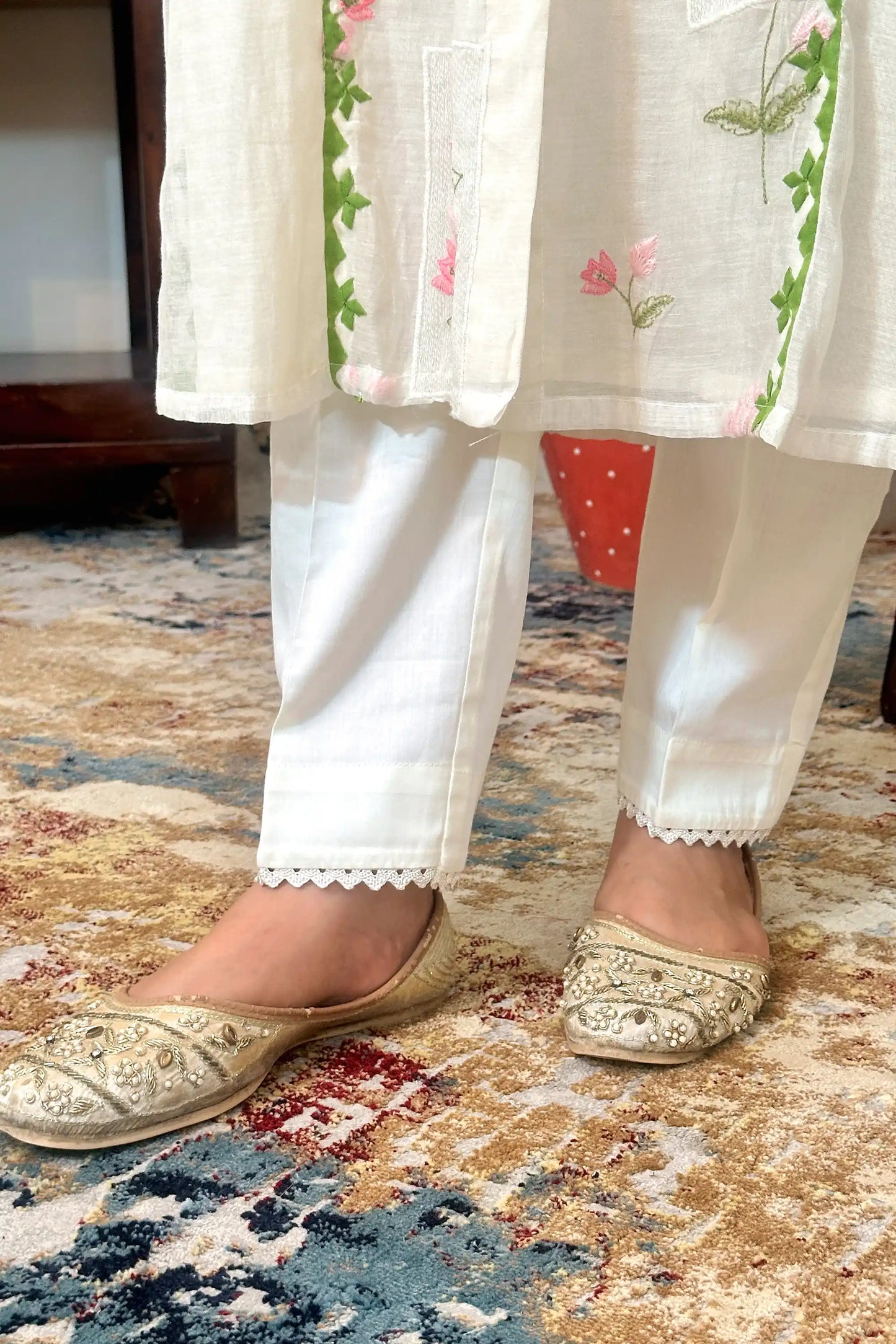 Close-up of the hem of the off-white chanderi kurta with pink floral embroidery and green applique trim, over off-white trousers with lace-edged hems.