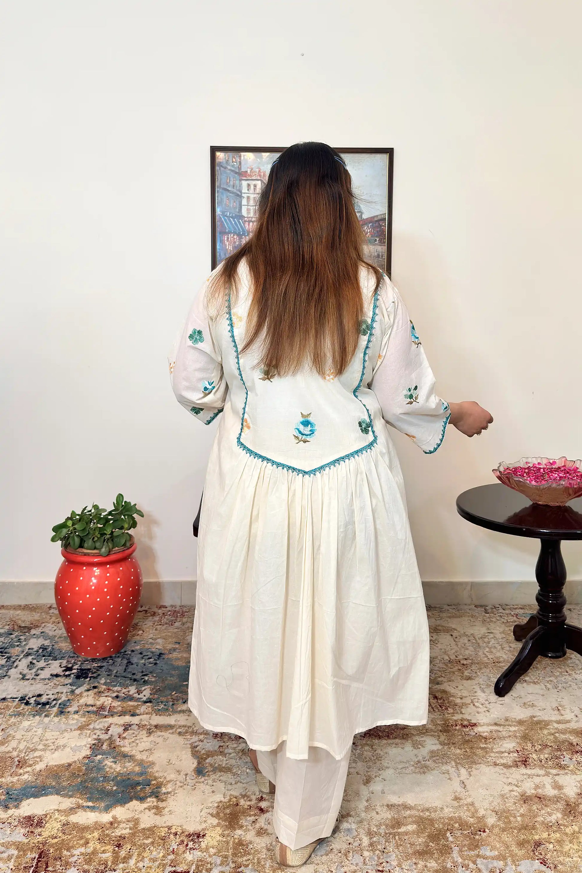 Back view of an off-white cotton frock kurta with multicolour floral resham embroidery on the yoke and sleeves, worn with off-white trousers.