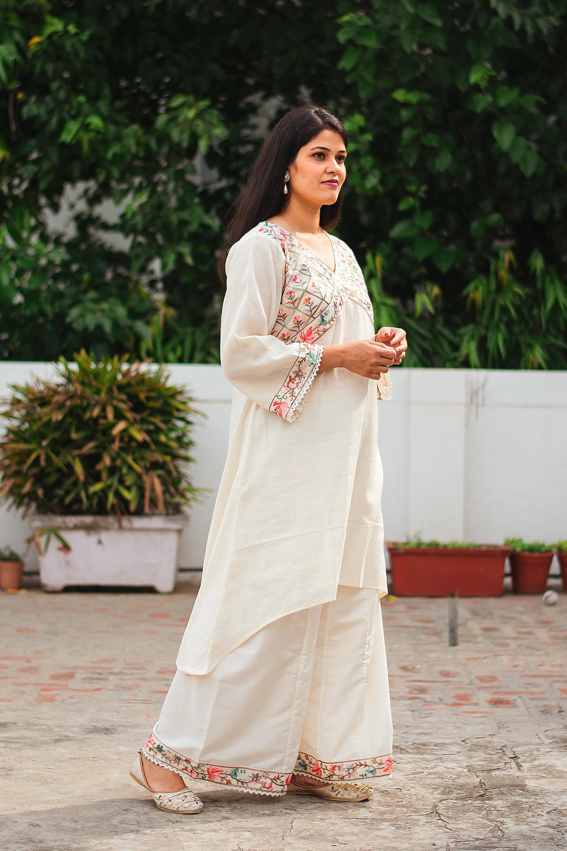 An Indian woman standing in a graceful side pose, showcasing the flowing silhouette and intricate details of an off-white asymmetric kurta