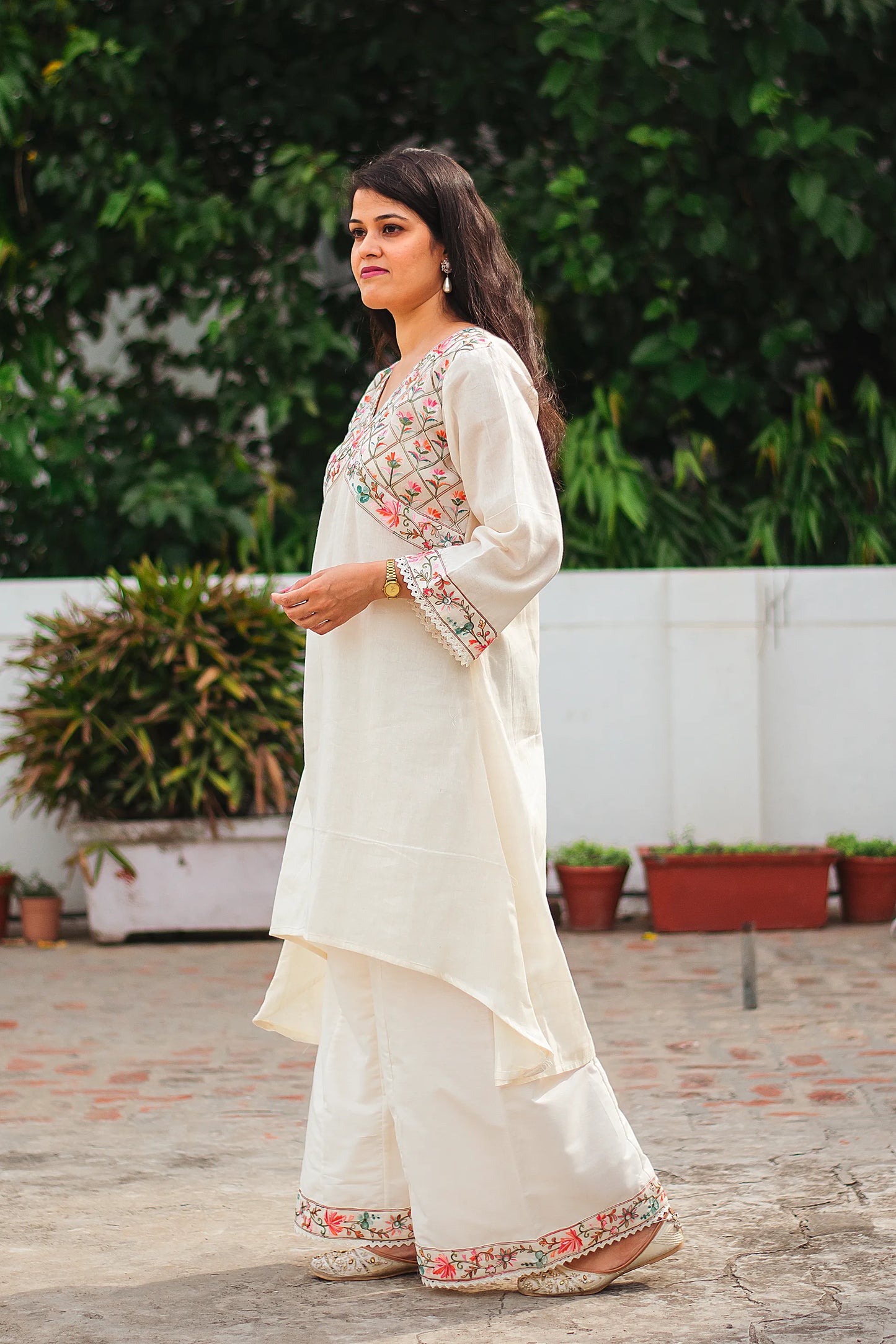 An Indian woman stands in a side profile highlighting the elegant drape of an off-white asymmetric kurta