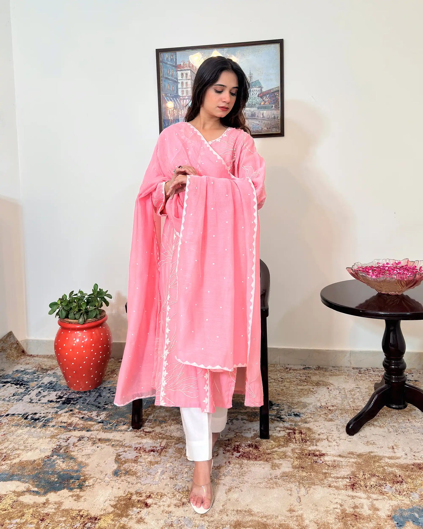 Model standing and arranging the dupatta with both hands near the chest, looking down, with the pink chanderi kalidar kurta and off-white cotton-silk trousers visible.