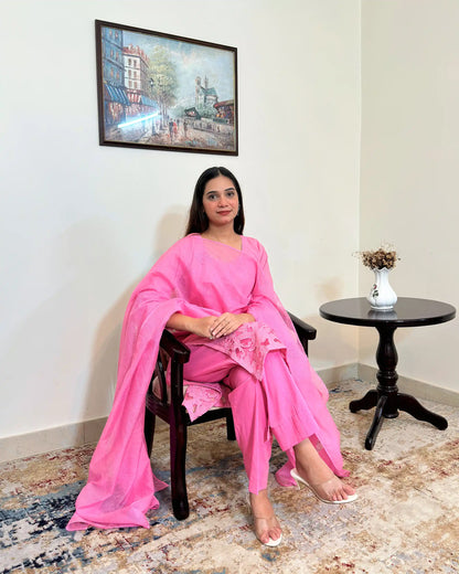 Model seated in a chair, hands folded, wearing the pink cotton straight kurta and sheer pink supernet dupatta, with the floral cutwork hem visible across the lap.