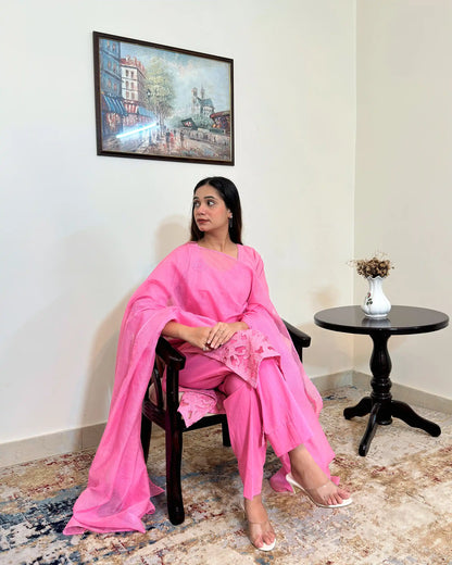 Model seated in a chair, looking to the side, wearing the pink cotton straight kurta and sheer pink supernet dupatta, with the floral cutwork hem visible across the lap.