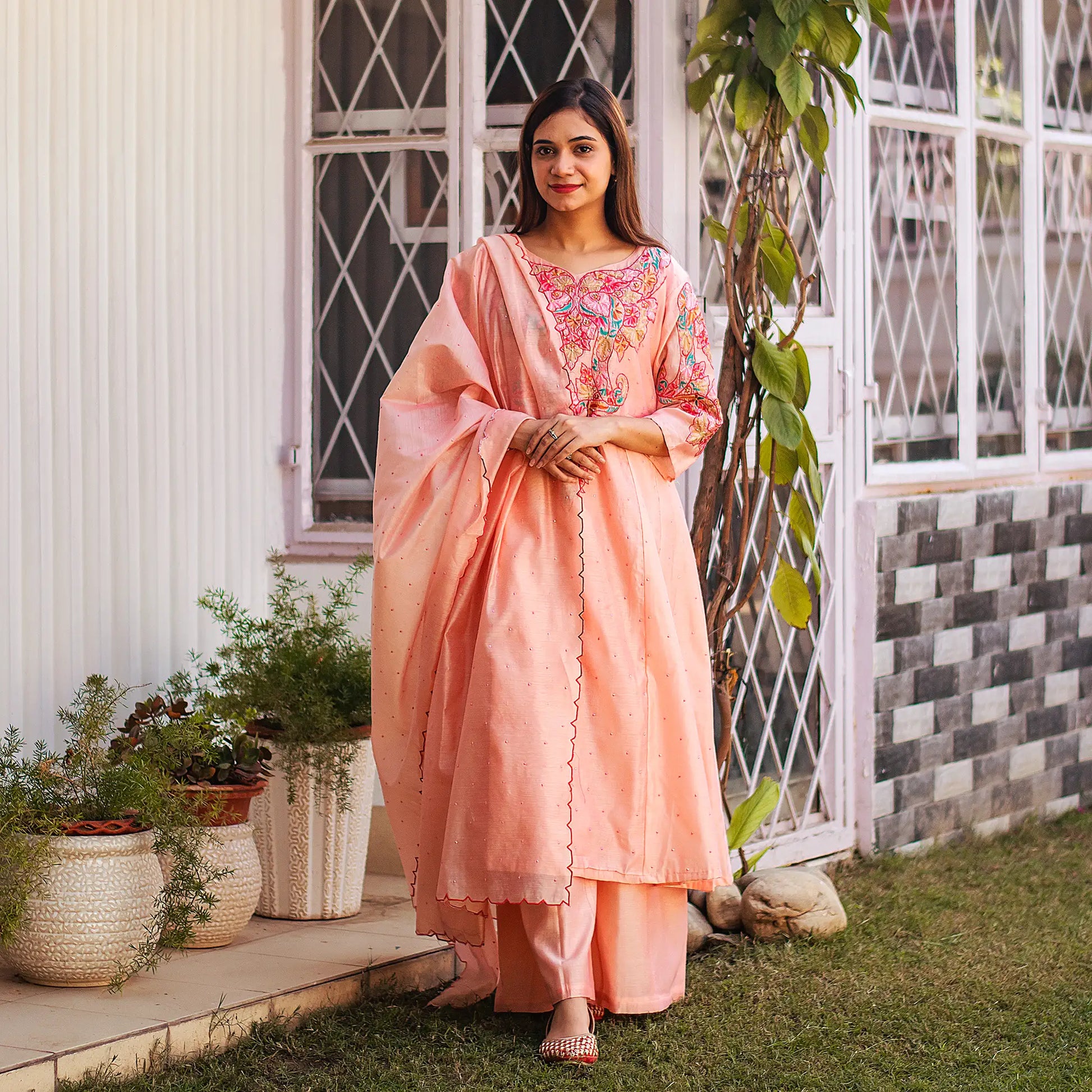  Indian woman dressed in a peach Chanderi kurta set with floral resham embroidery and pink sequins, holding a dupatta.