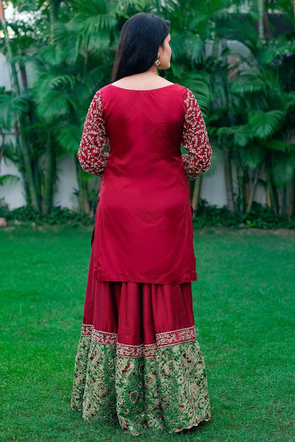 A woman wearing a maroon silk Gharara with a matching tissue dupatta draped over her head and shoulders.