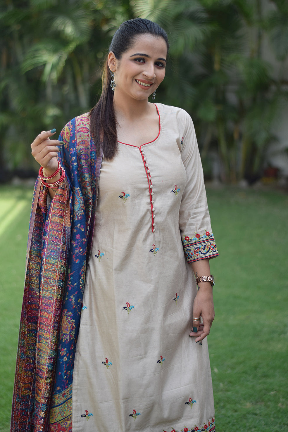 A woman poses wearing a beige kurta with a blue silk jamawar dupatta draped over her arms.