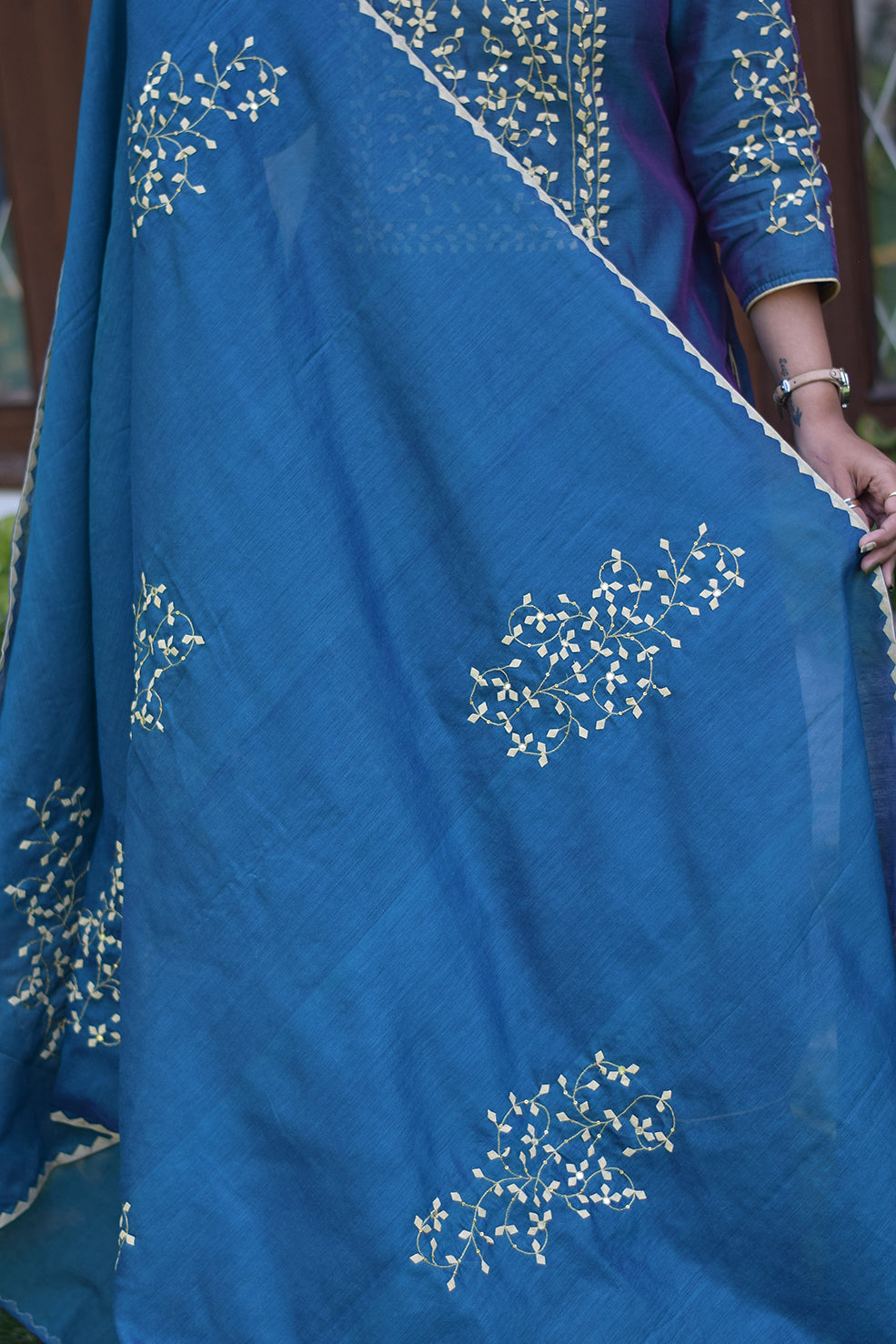 A solo Indian woman standing tall in a blue applique work suit with her arms folded.