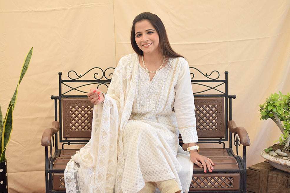 A woman sitting gracefully, wearing a white georgette chikankari kurta with intricate kikri and kamdani embroidery, paired with a matching dupatta and golden trousers