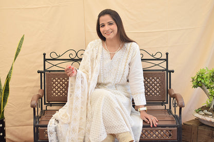 A woman sitting gracefully, wearing a white georgette chikankari kurta with intricate kikri and kamdani embroidery, paired with a matching dupatta and golden trousers
