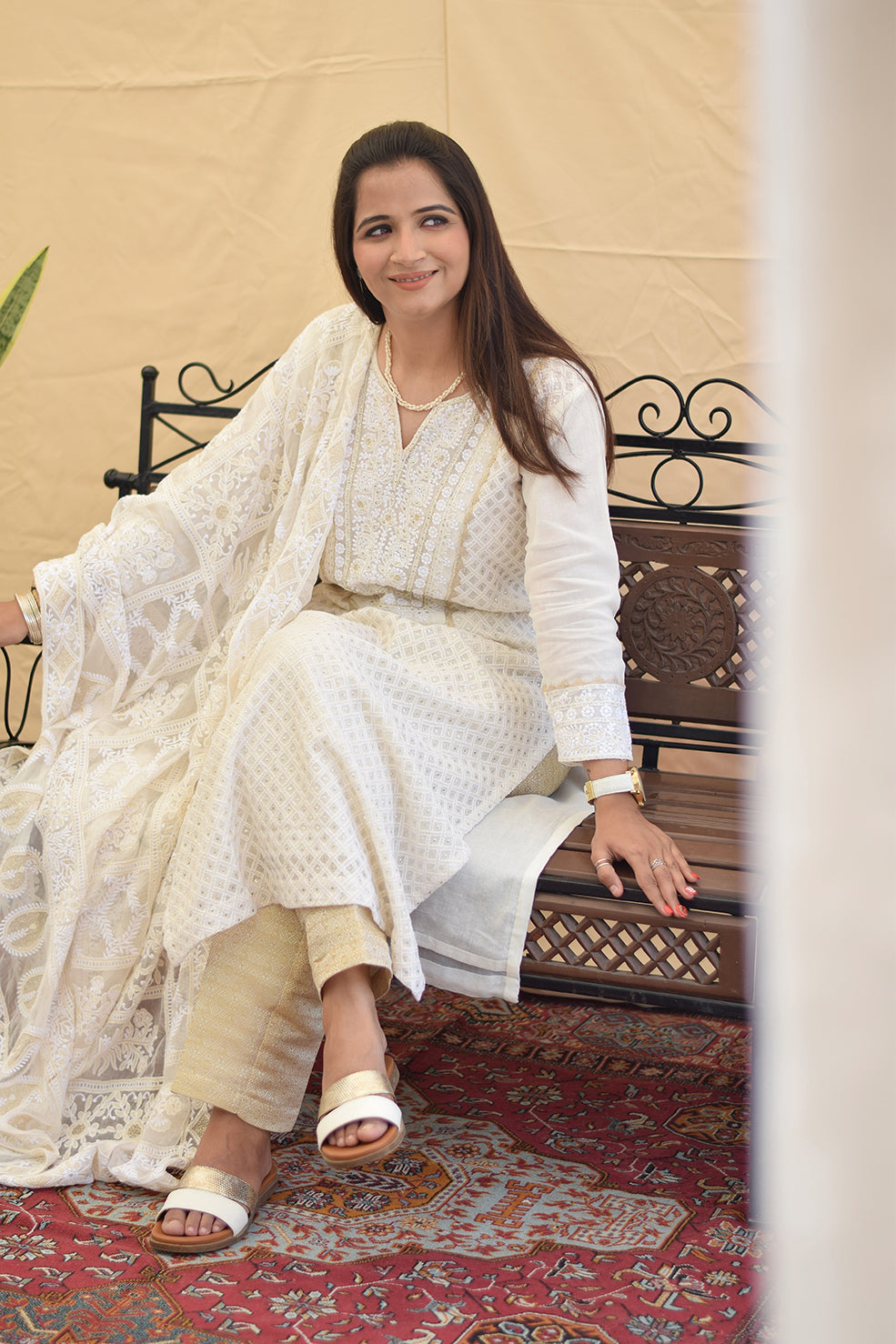 A woman sitting on a bench, wearing a white georgette chikankari kurta with intricate kikri and kamdani embroidery, paired with a matching dupatta and golden trousers.