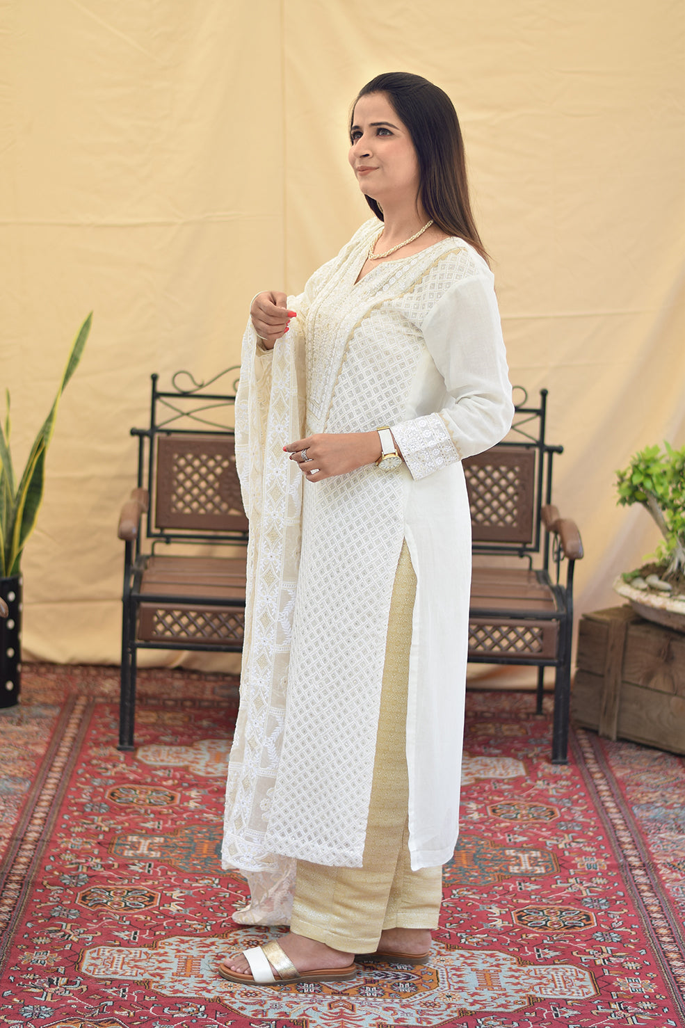 A woman in a side pose, wearing a white georgette chikankari kurta with intricate kikri and kamdani embroidery, paired with a matching dupatta and golden trousers.